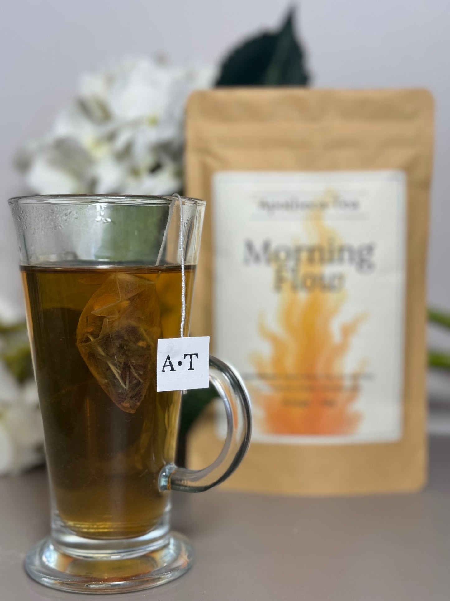 Glass mug with tea and a tea bag labeled 'A-T' next to a package of 'Morning Flow' tea.