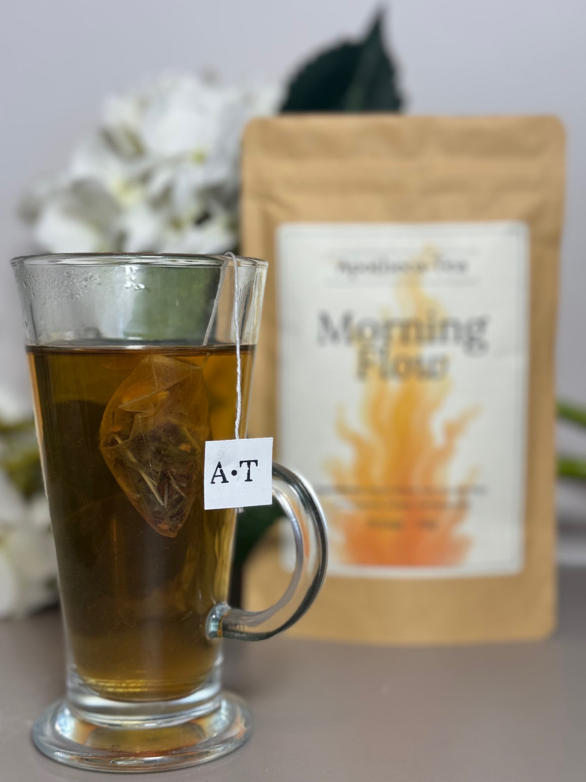 Glass mug with tea and a tea bag labeled 'A-T' next to a package of 'Morning Flow' tea.