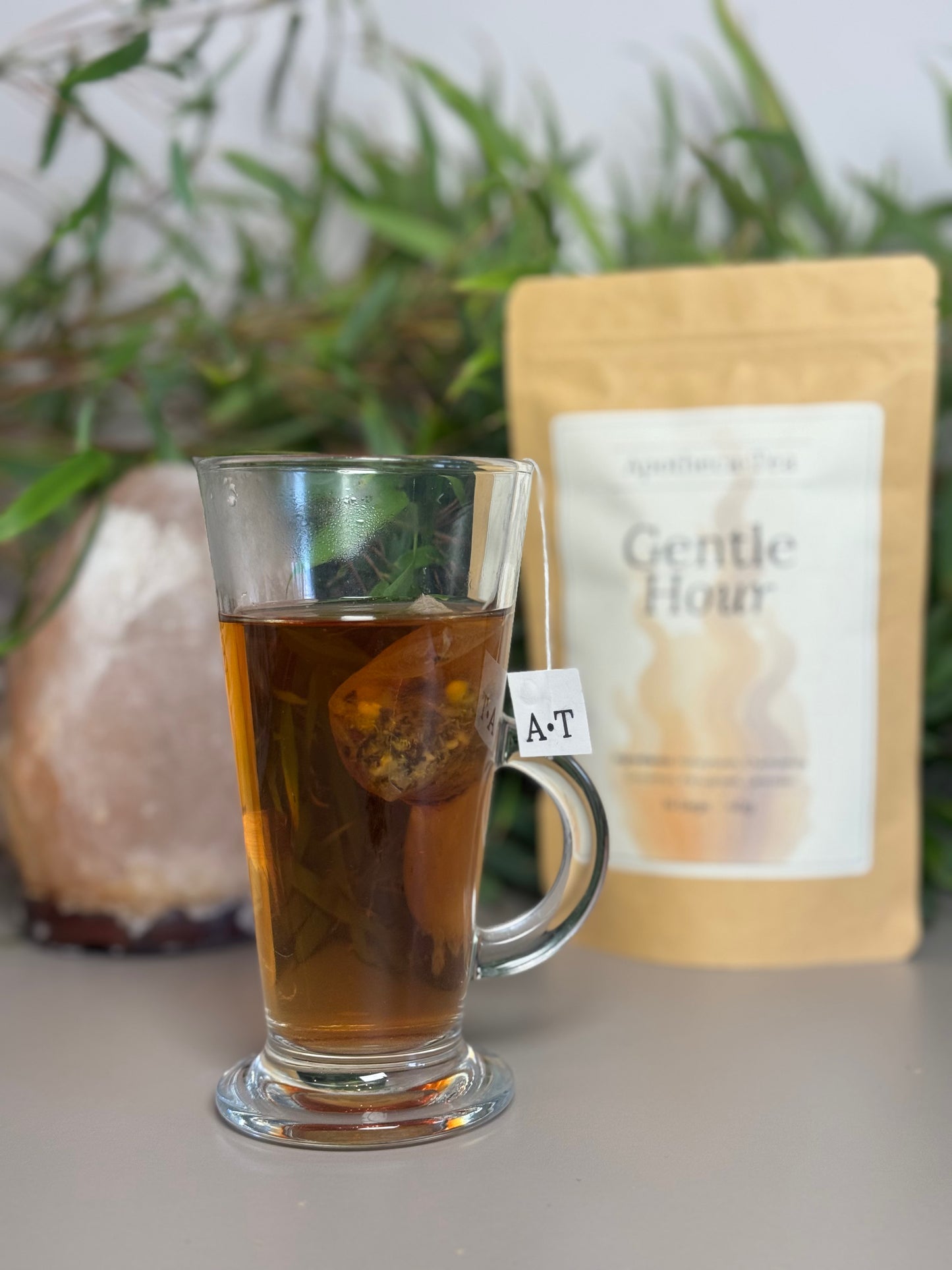 A portrait shot of apotheca tea gentle hour blend in a glass mug displaying the color of the tea and tea bag. gentle hour packet is seen in the background with greenery and salt lamp.